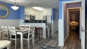 A modern kitchen with white cabinets, a stainless steel refrigerator, and a dining area with white chairs and a table. - Dolphin Cove 3A - Oceanfront Condominium in Garden City Beach (Garden City)