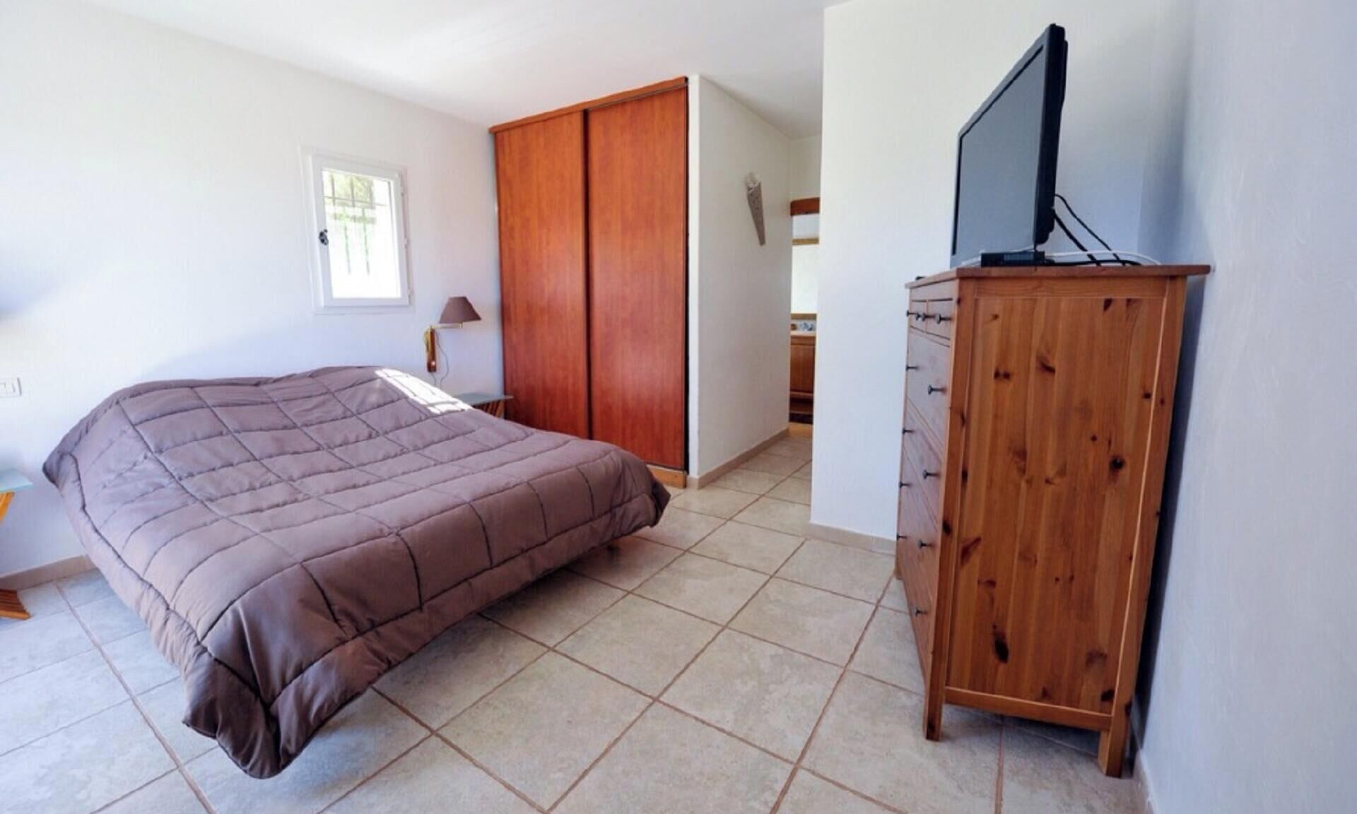 Une chambre à coucher avec un lit, une armoire en bois, un téléviseur posé sur un meuble et une fenêtre.