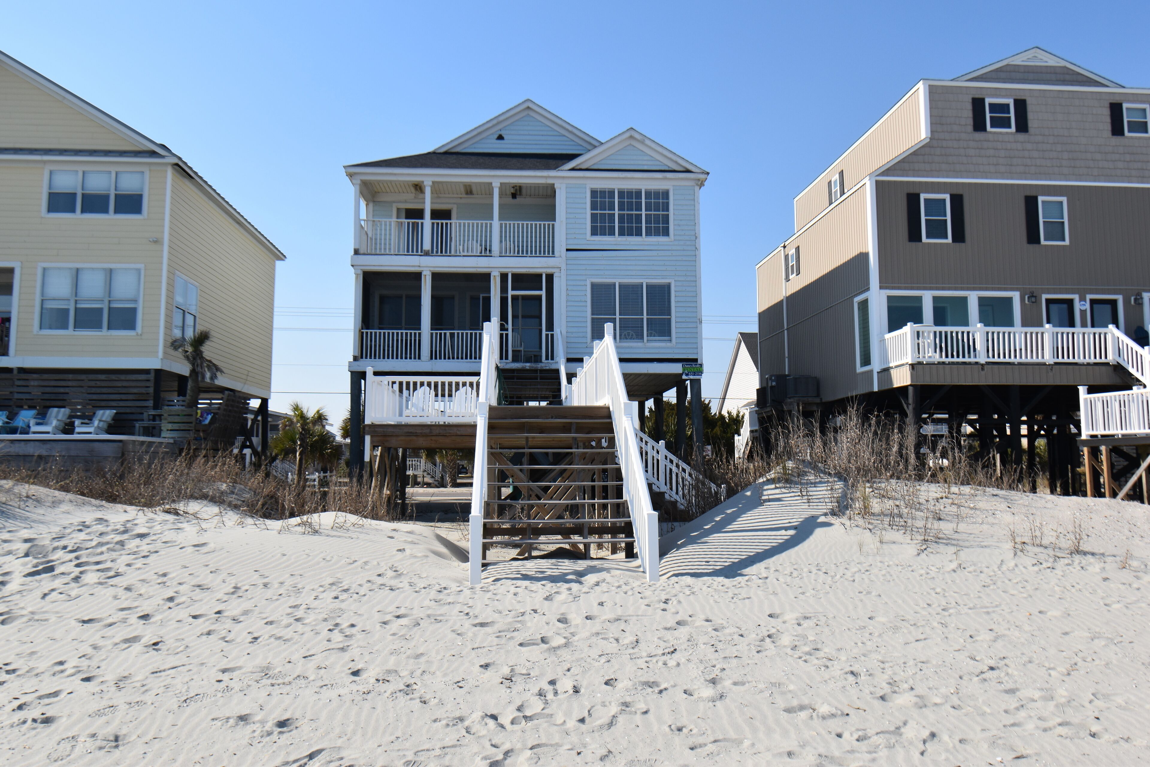 A beachfront property with a multi-level house, a staircase leading to a deck, and a sandy beach in the foreground.