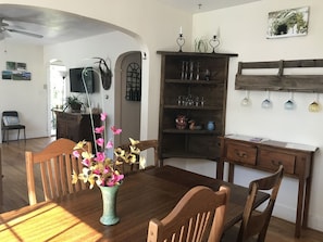 Uma área de refeições com mesa de madeira, cadeiras, um vaso com flores, uma estante e uma prateleira na parede com objetos decorativos.
