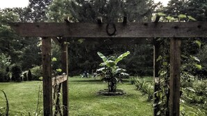 A wooden pergola structure with a circular stone base, surrounded by a well-maintained grassy area and various plants. - Coburn On the Creek - Flyfish, Tube , Grill, Relax- PSU Football/Camp Woodward (Coburn)