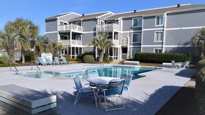 A pool area with lounge chairs and a table, surrounded by a multi-story building with balconies. - Sea Cloisters I 101B - Oceanfront Condominium in Surfside Beach (Surfside Beach)