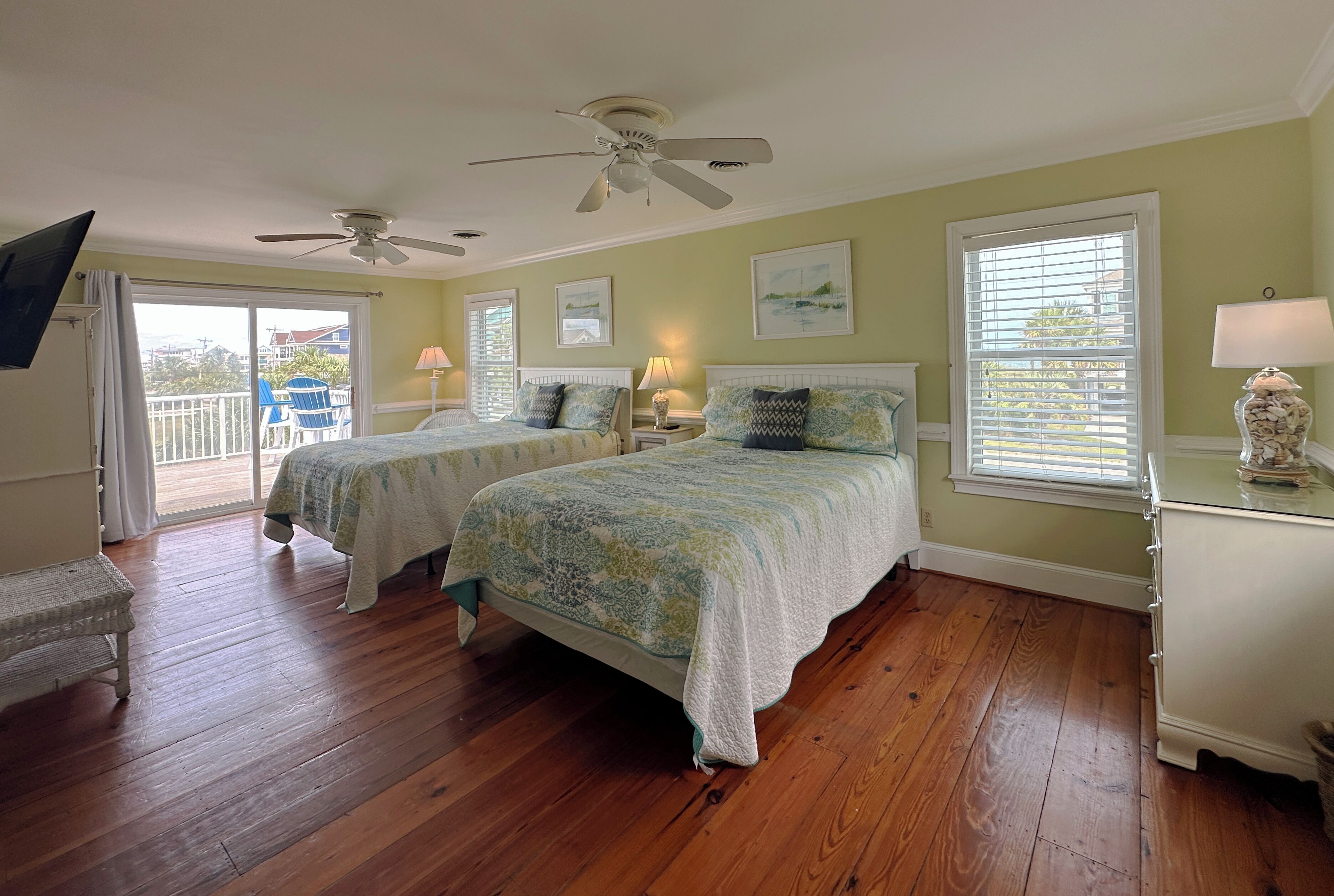 A room with two beds, a ceiling fan, a balcony with a chair, and a view of the outside.