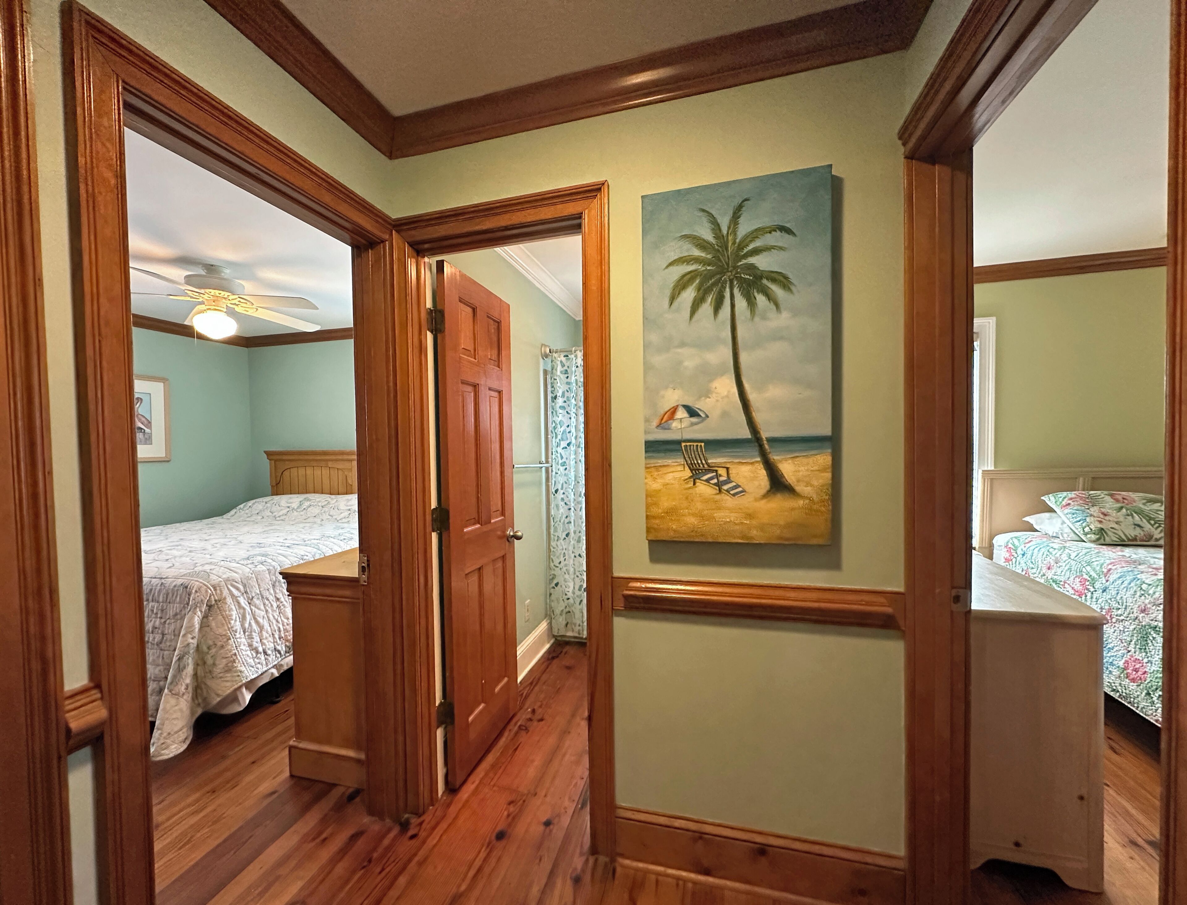 A hallway with wooden floors and doors leading to a bedroom with a bed and a painting of a beach scene.