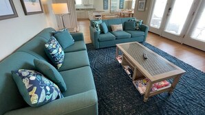 A living room with a sofa set, a coffee table, and a rug. There is a kitchen in the background.