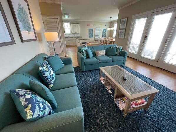 A living room with a sofa set, a coffee table, and a rug. There is a kitchen in the background.