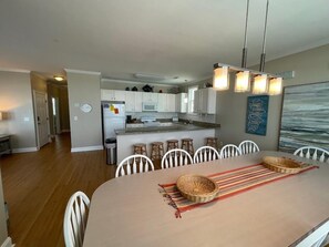 A dining area with a large table, white chairs, and a kitchen in the background.