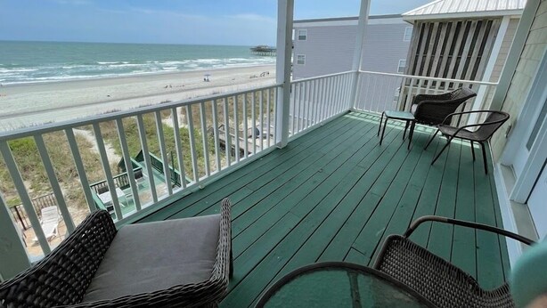 A covered balcony with wicker furniture, a view of the beach, and a house in the background.