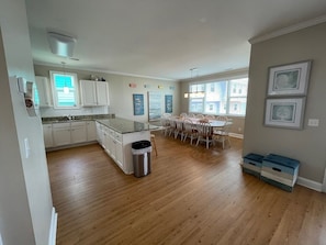 A spacious kitchen with wooden flooring, white cabinets, a dining area with a table and chairs, and a trash bin.