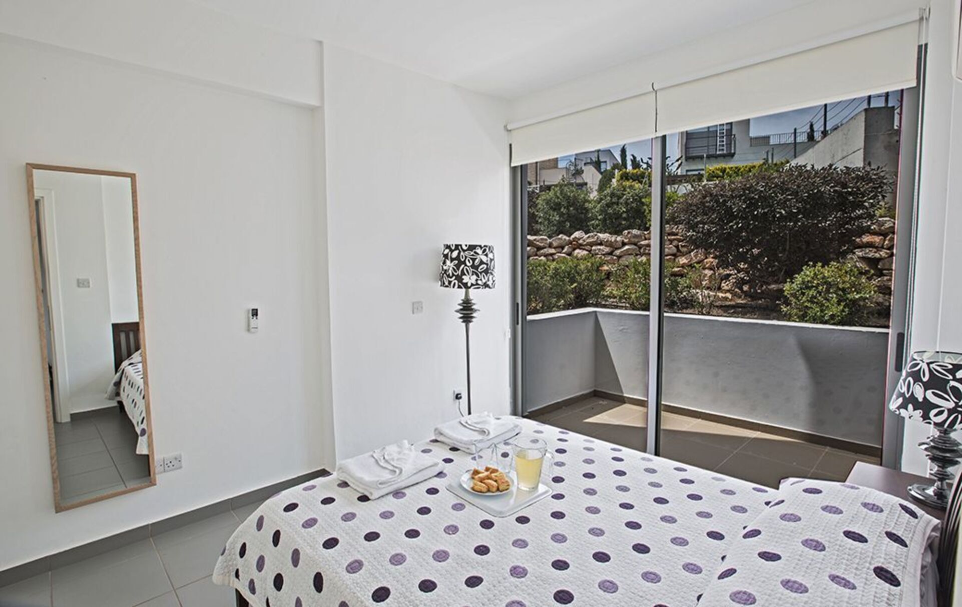 Ein Schlafzimmer mit einem Bett, einem Nachttisch mit Keksen und einem Glas Wasser, einer Stehlampe und einem großen Fenster mit Blick auf Grünflächen und Gebäude.