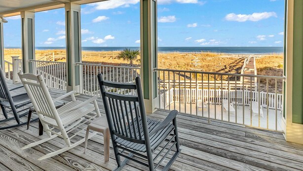 Una terraza techada con piso de madera y barandas blancas, con vista a una playa con dunas de arena y un cielo azul despejado.