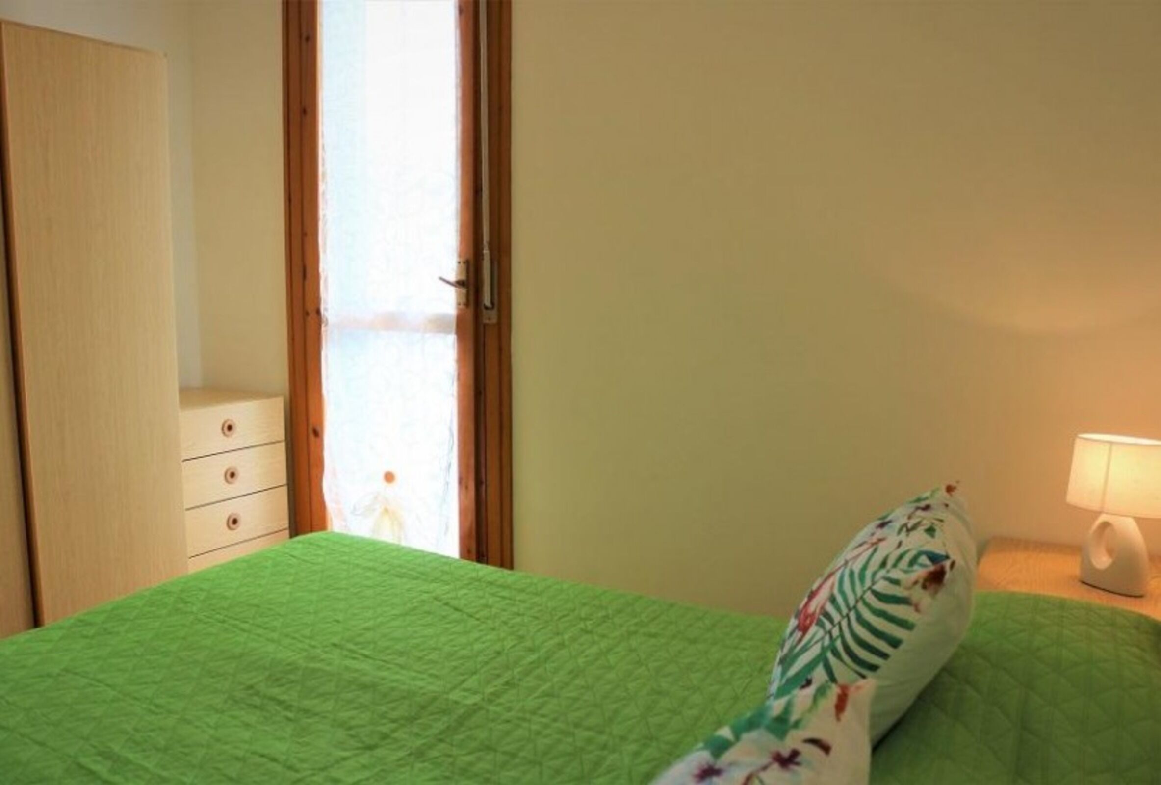 Una habitación con colcha verde, un armario de madera, una mesita de noche con lámpara y una ventana con cortinas.
