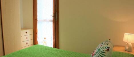 Una habitación con colcha verde, un armario de madera, una mesita de noche con lámpara y una ventana con cortinas.