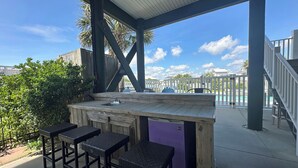 An outdoor bar area with a wooden counter, a sink, and three black wicker stools. - Buckeye Heaven - Second Row in Garden City Beach (Garden City Beach)