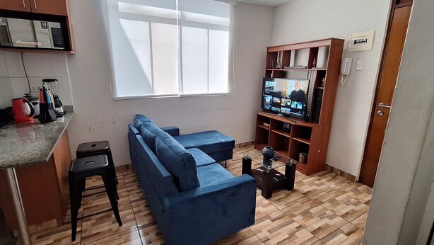 A compact living space with a kitchenette, a blue sofa, a television, and a wooden shelf unit. - LIMA FLAT 1 1D (10 MIN FROM MIRAFLORES) (Lima)