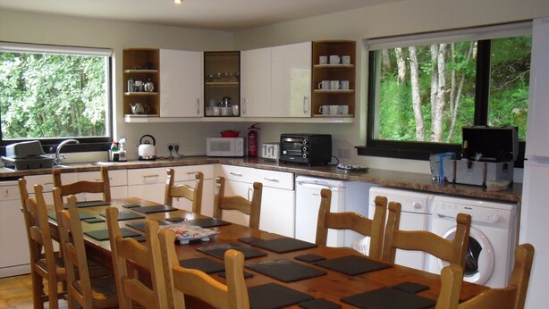 A modern kitchen with white cabinets, a wooden dining table with chairs, a built-in oven, and a window with a view of trees. - Comfortable property with ample space and views of the 5 sisters of Kintail (Glenshiel)