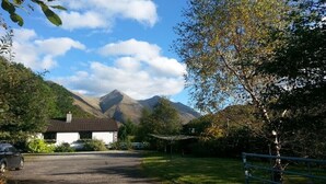 A rural property with a gravel driveway leading to a white building with a tiled roof, surrounded by lush greenery and mountains in the background. - Comfortable property with ample space and views of the 5 sisters of Kintail (Glenshiel)