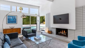 A modern living room with a sofa, coffee table, fireplace, TV, and large windows. - Desert Eden – Midcentury Pool Oasis (Palm Springs)
