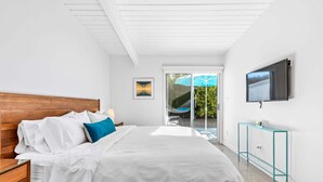 A bedroom with a wooden bed, white bedding, a TV mounted on the wall, and a glass door leading to an outdoor area. - Desert Eden – Midcentury Pool Oasis (Palm Springs)