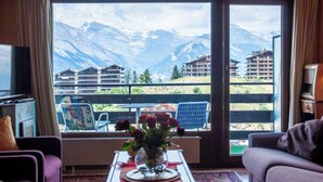 Een kamer met een groot raam en uitzicht op bergen, een tafel met een bloemenvaas, en een bank.