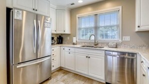 A kitchen with white cabinets, stainless steel appliances, a window with blinds, and a tiled floor. - The Taylor at Ships Watch in Duck (Duck)