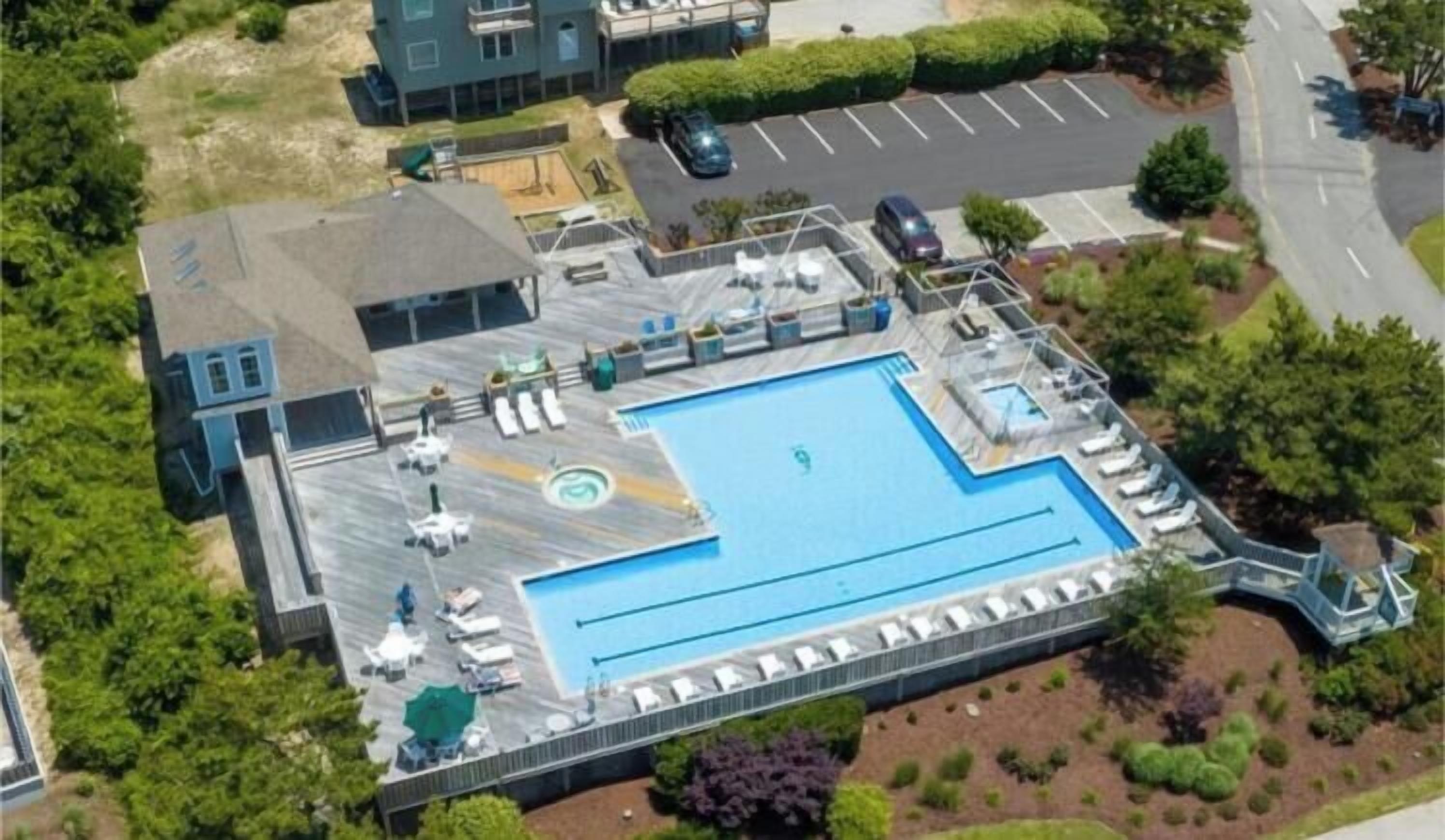 A swimming pool with lounge chairs and a covered area, surrounded by greenery and a parking lot.