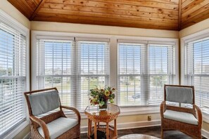 A sunroom with large windows, wicker chairs, a small table with a vase, and a wooden ceiling. - The Taylor at Ships Watch in Duck (Duck)