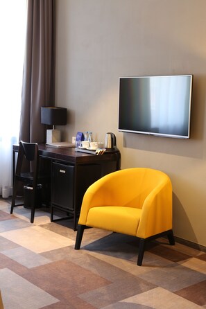 Habitación de hotel con escritorio, un sillón amarillo, televisor de pantalla plana, lámpara y cafetera.