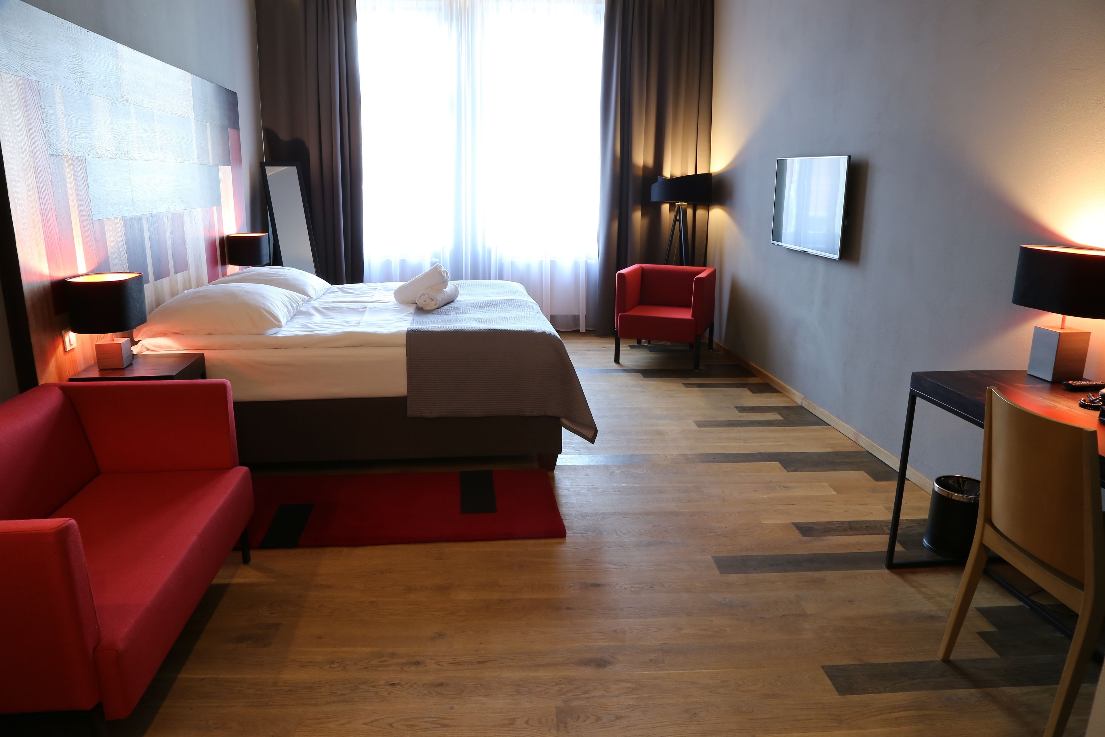 A hotel room with a large bed, a red armchair, a small table with a lamp, and a modern wall-mounted TV.