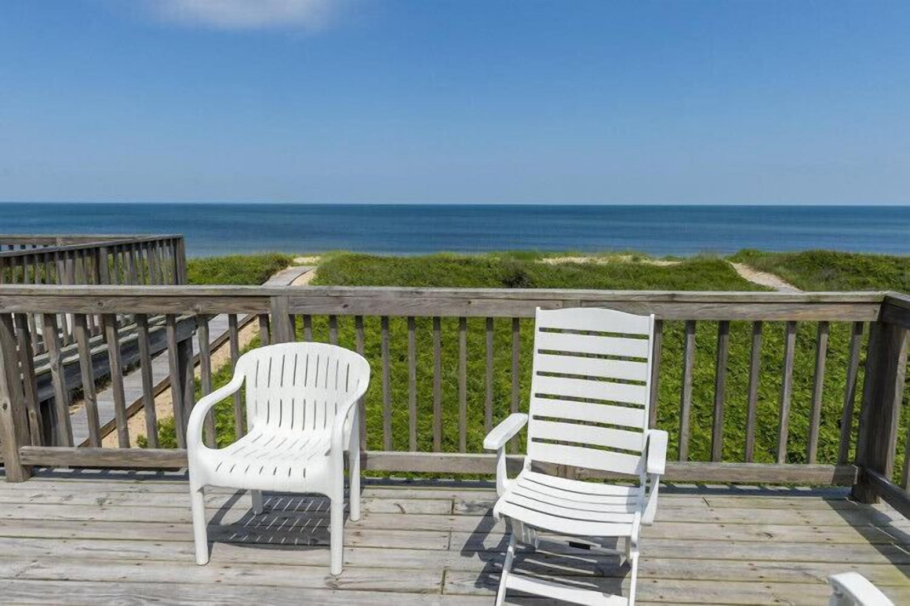 A wooden deck with two white chairs facing a beach view.