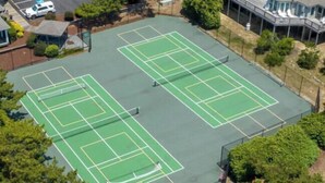 En vy från ovan av ett tennisanläggningsområde med flera banor, omgivet av träd och byggnader.