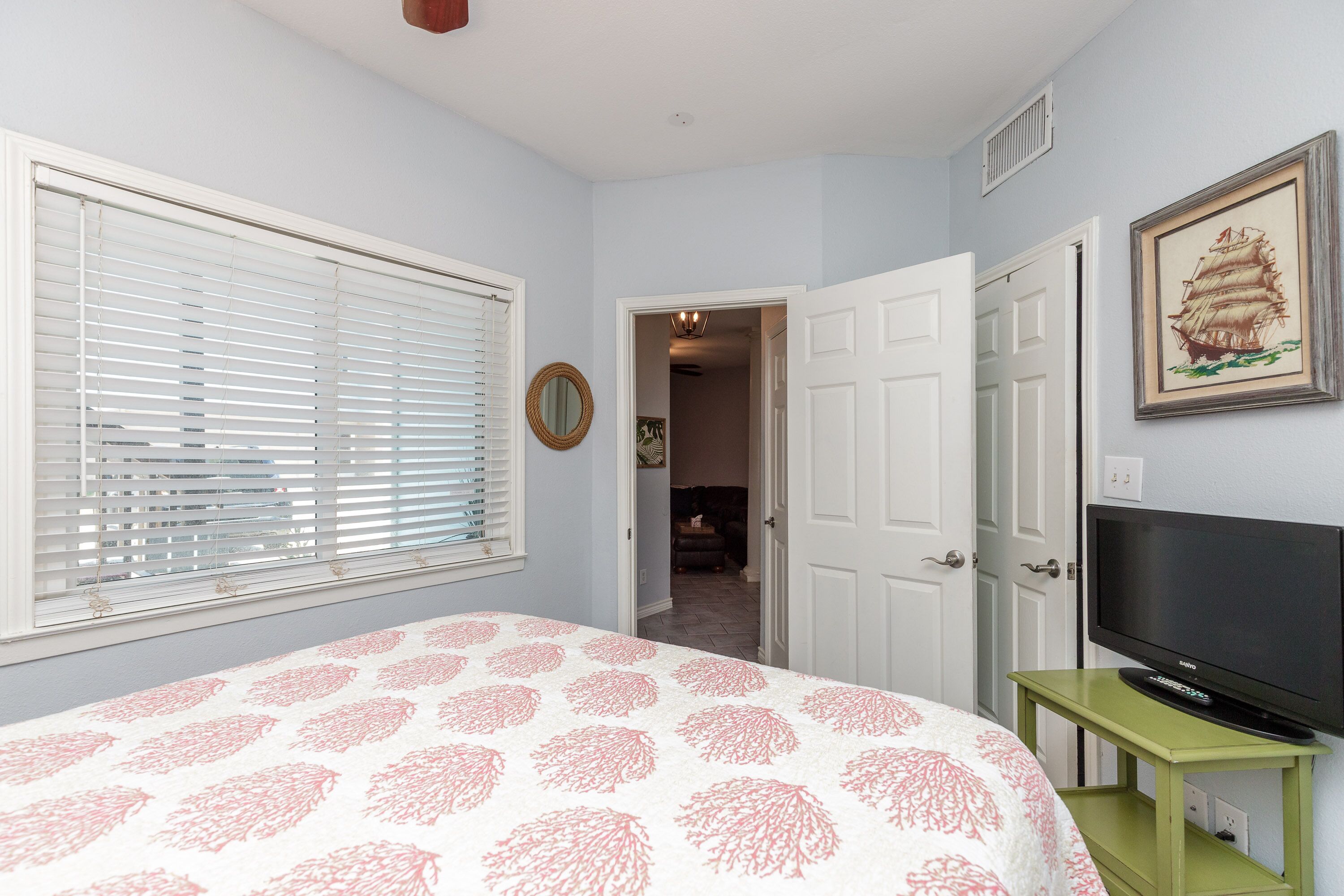 A bedroom with a bed, a television on a stand, a door leading to another room, and a window with blinds.