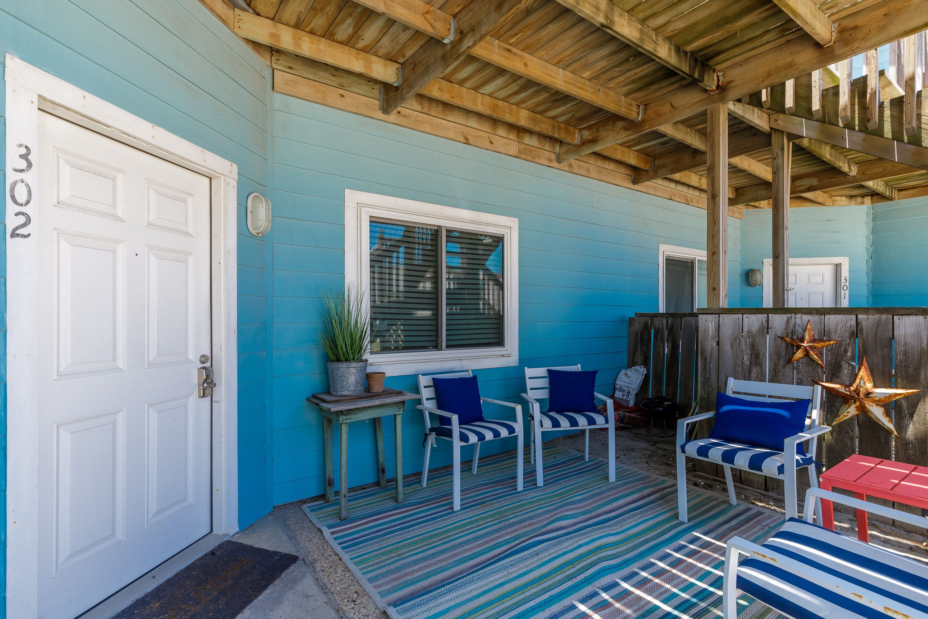 A covered porch with a blue exterior, a white door marked "302", two white chairs with blue cushions, a small table with a potted plant, and a red table.