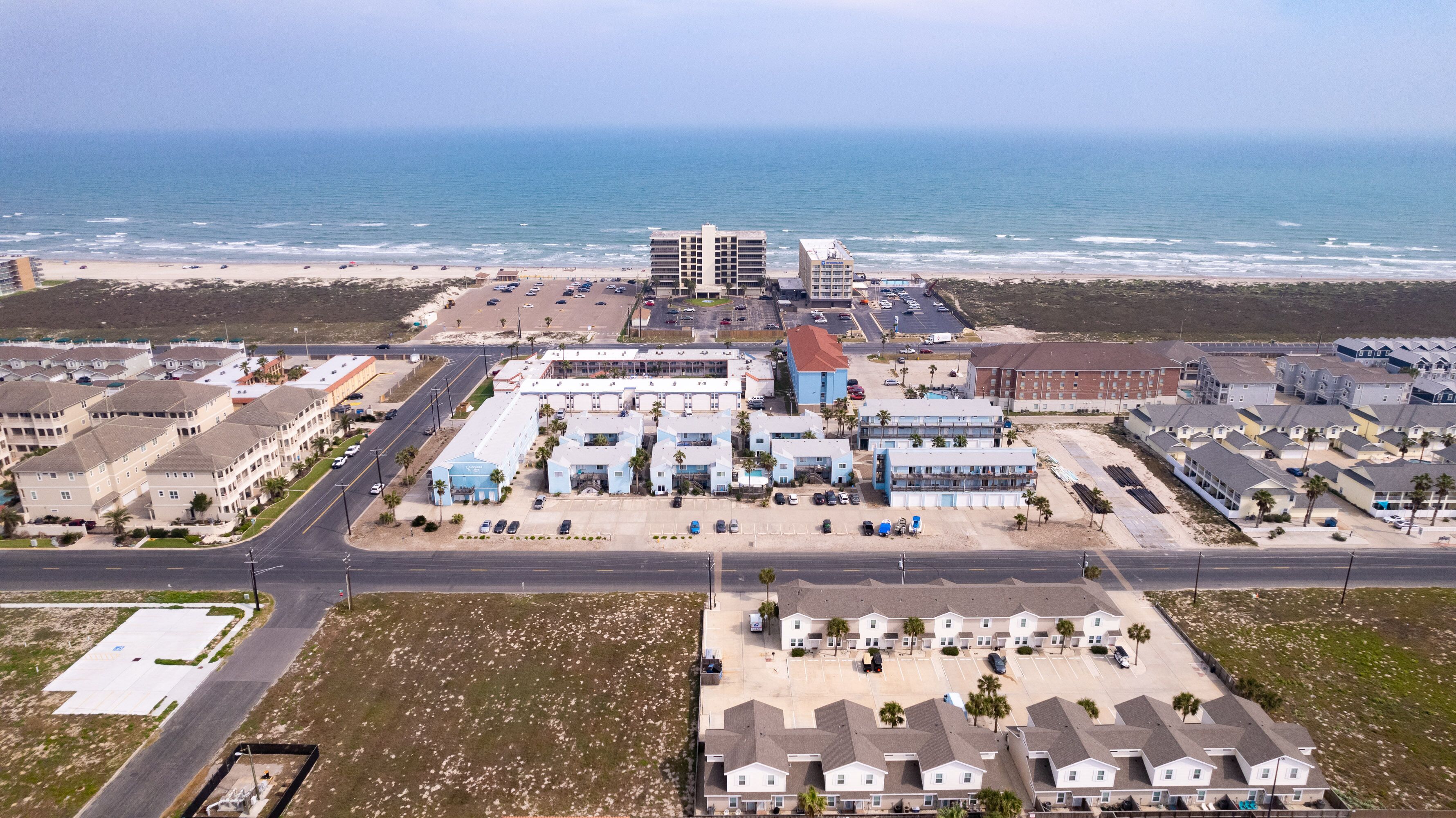 An aerial view of a coastal hotel complex with multiple buildings, a parking lot, and a beach in the background.