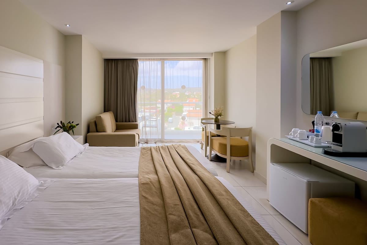 Une chambre d’hôtel avec deux lits, un bureau avec une machine à café, une chaise, une vue sur la ville grâce à de grandes fenêtres et un balcon.