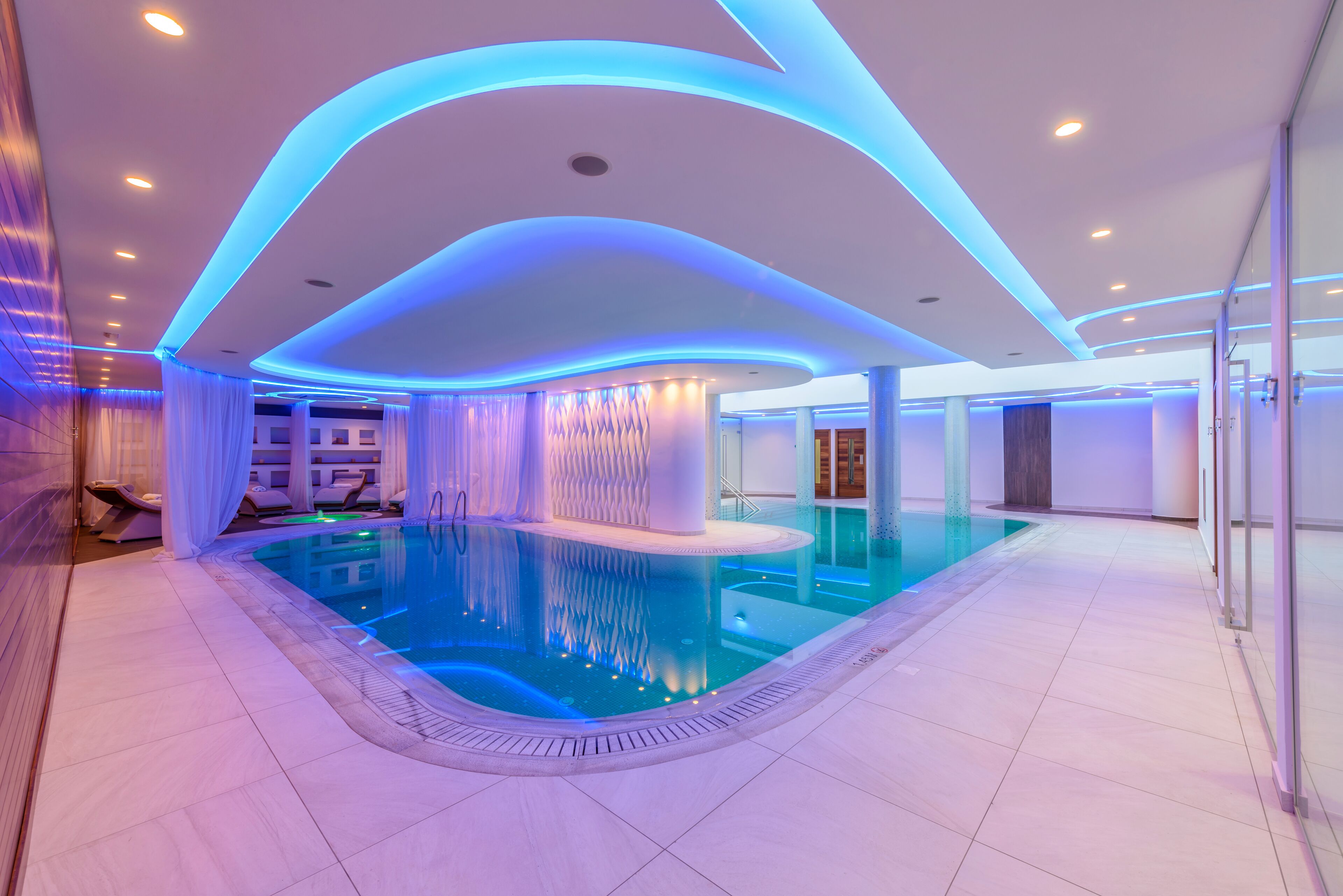 Piscina interior moderna con iluminación azul, una fuente central y una zona de descanso con cortinas blancas.