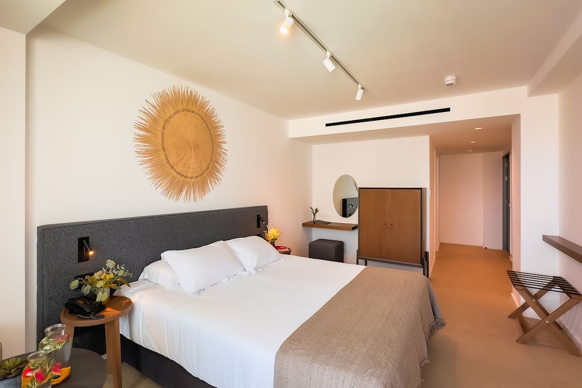 Une chambre d’hôtel moderne avec un grand lit, un élément de décoration circulaire au mur, une table de chevet avec un téléphone et des fleurs, et une vue sur le couloir.