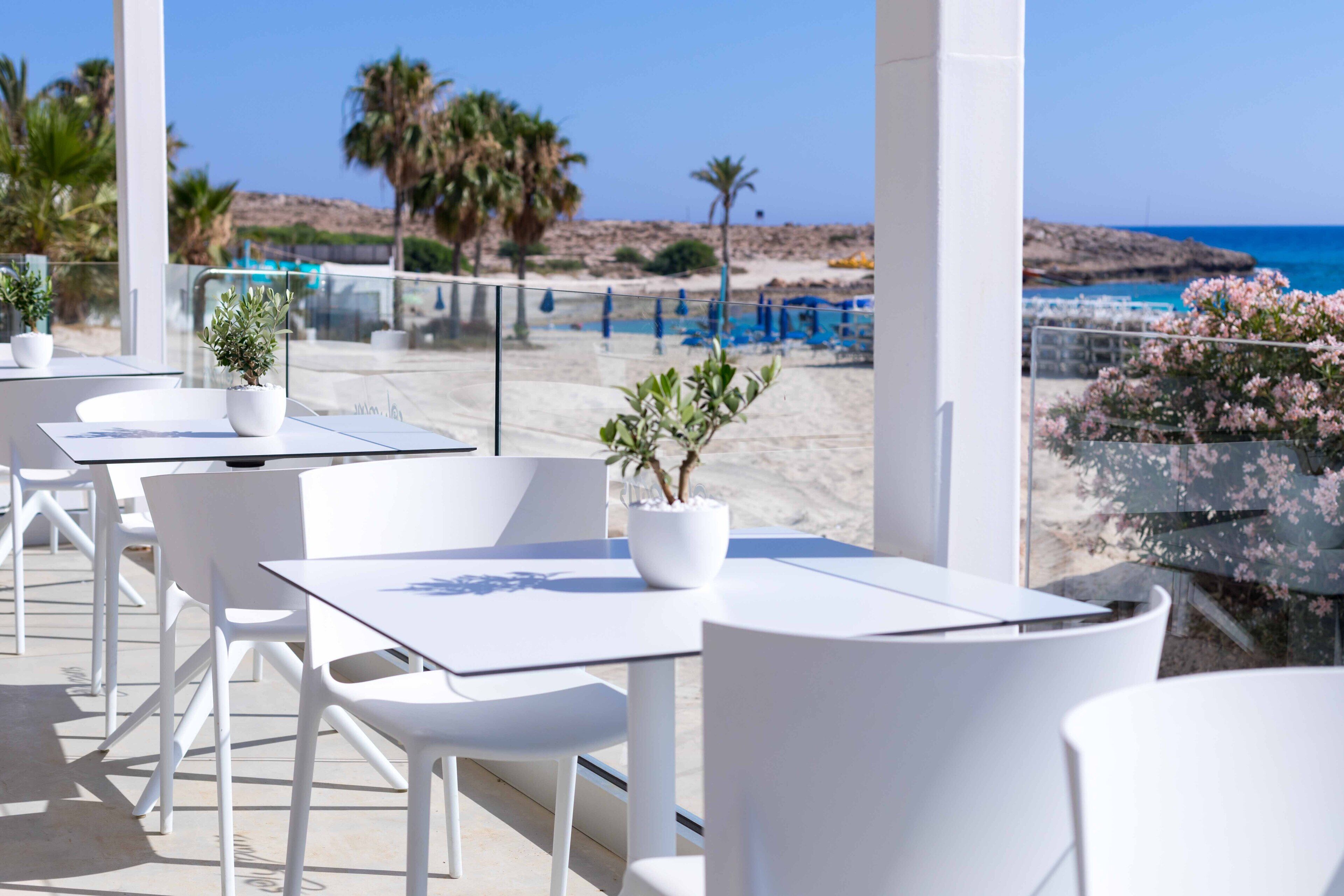 Un espace extérieur aménagé avec des tables et des chaises blanches, des plantes en pot et une vue sur la plage et l’océan.