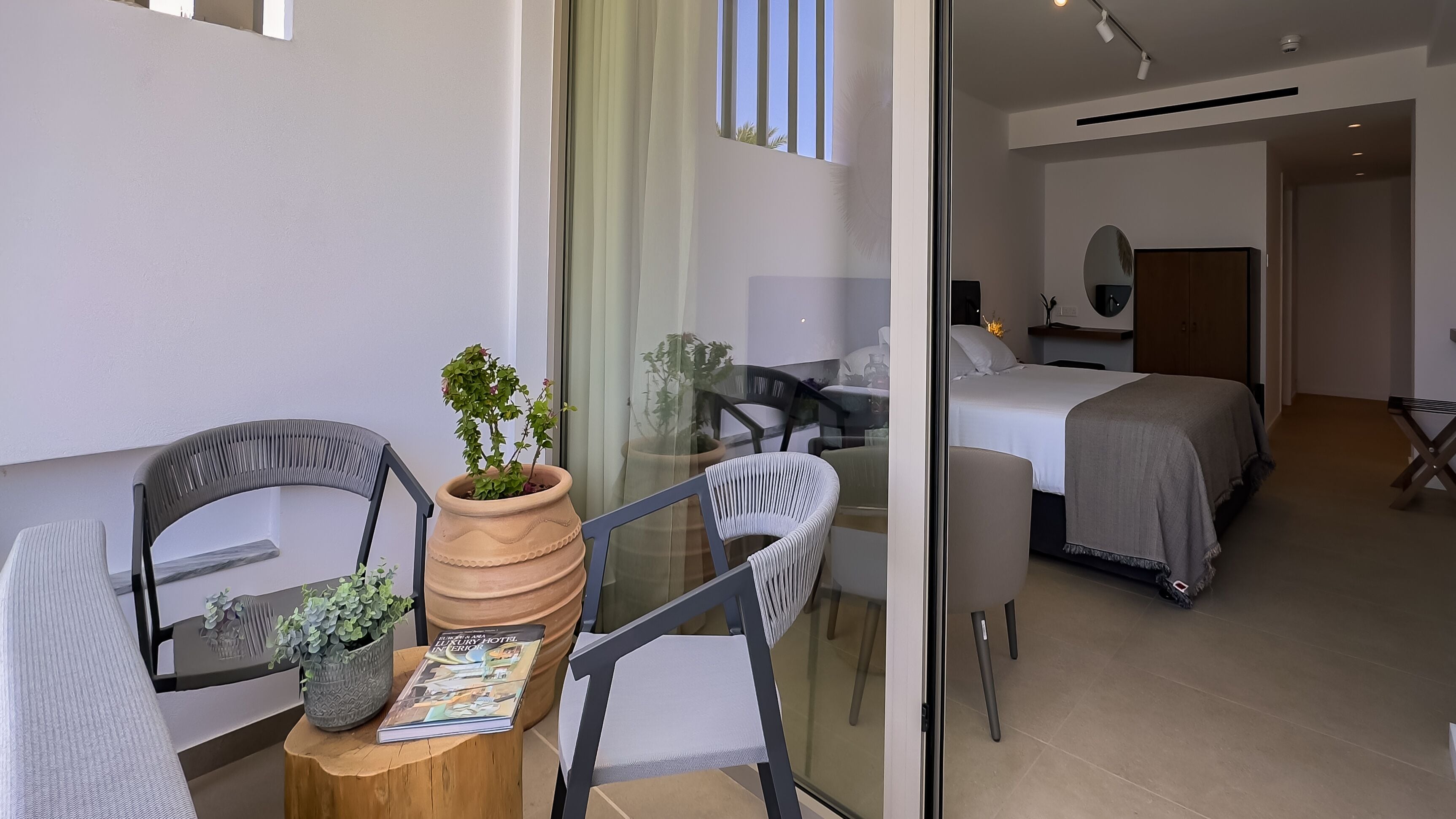 A modern hotel room with a glass partition, a dining area with a table and chairs, and a bed with a white bedspread.
