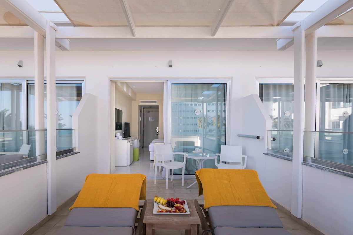 Une chambre d’hôtel moderne avec un balcon, une table à manger avec des fruits et un canapé sur lequel est posée une serviette jaune.