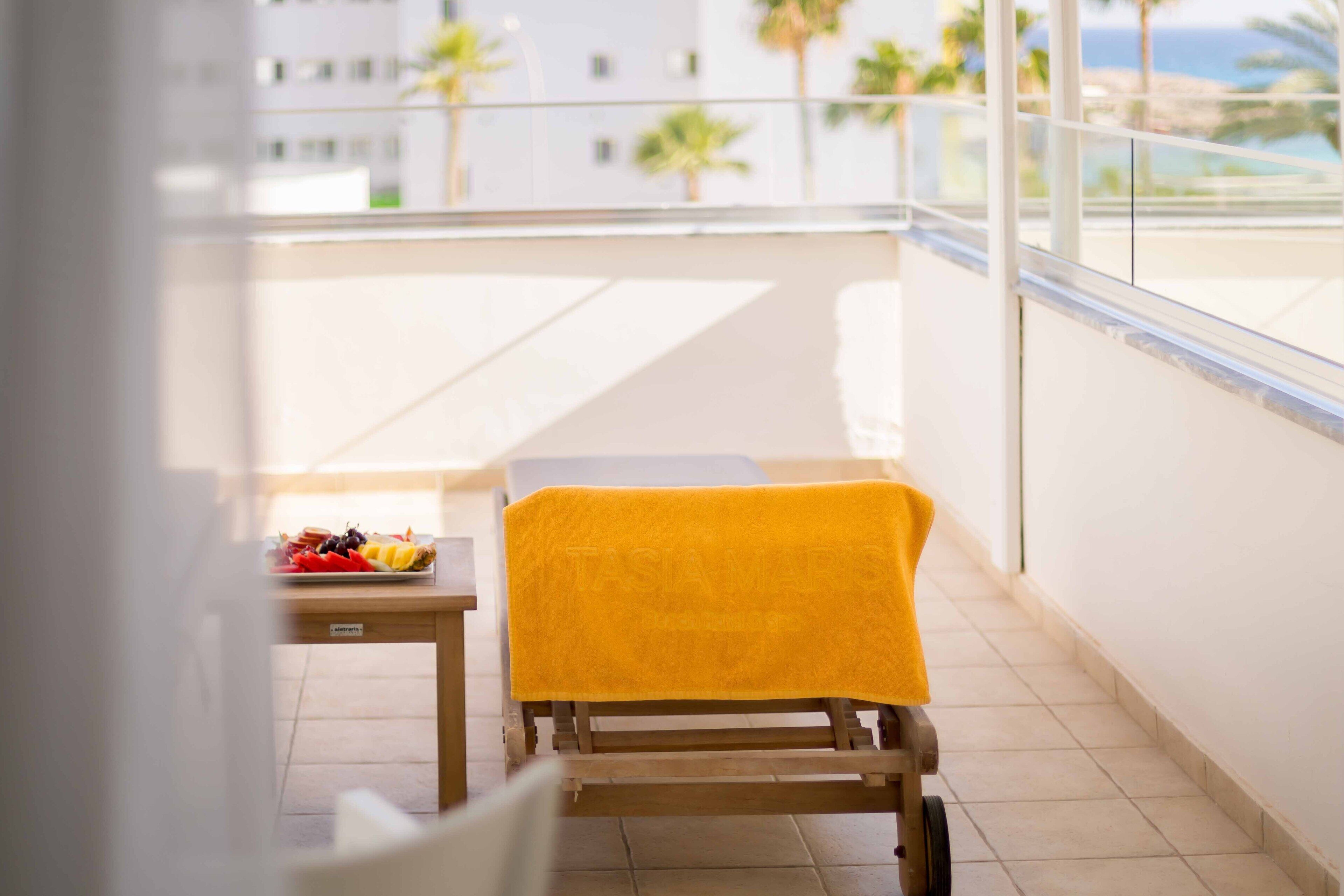 A balcony with a wooden chair covered by a yellow towel, a small table with a fruit platter, and a view of a building and palm trees.