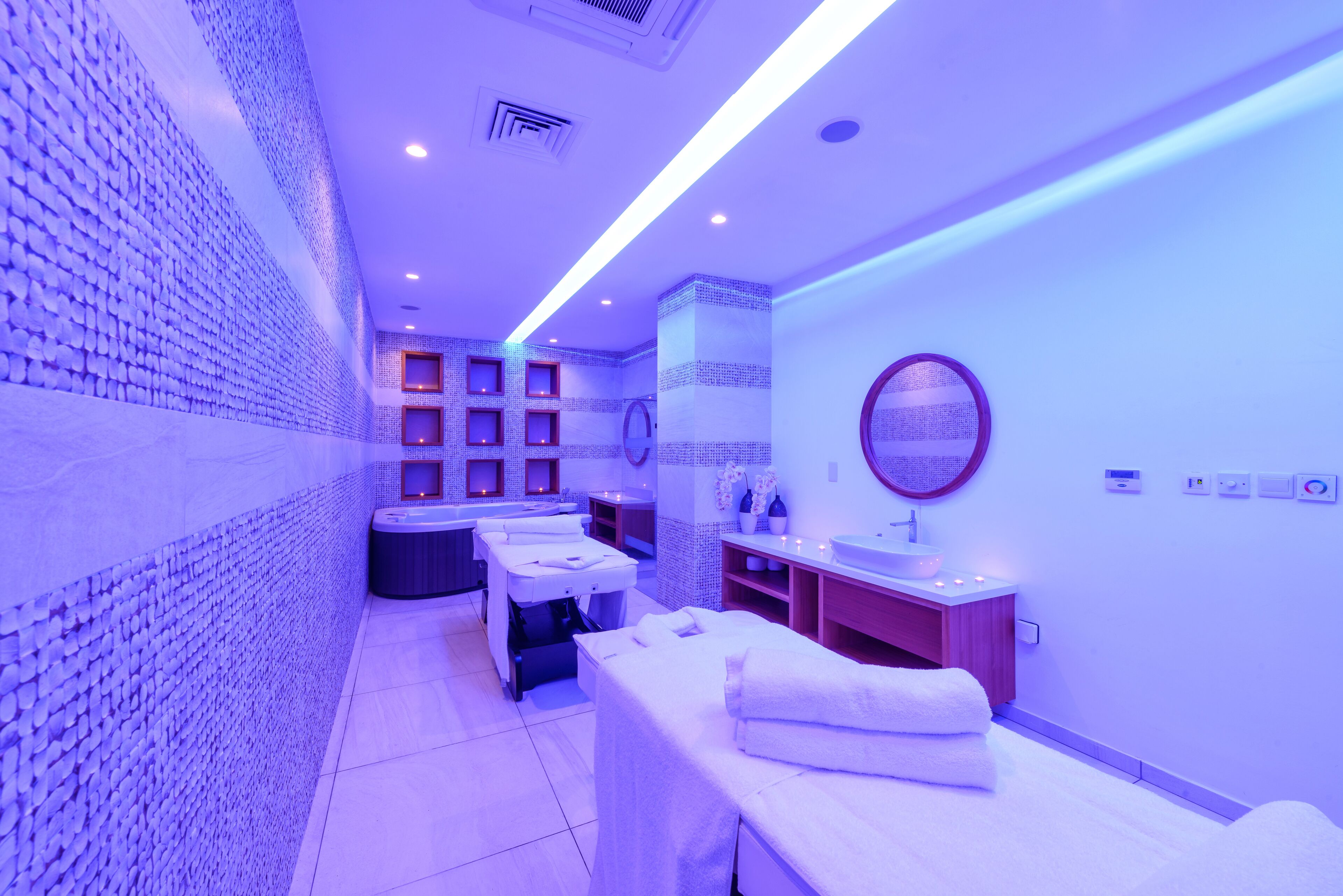 A spa room with a tiled wall, a massage table, a sink, and a mirror.