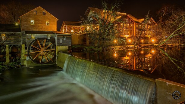 A historic building with a large water wheel, wooden exterior, and illuminated windows at night. - Porch w/ swing & hot tub, sounds of stream, fireplace, pet & family friendly! (Pigeon Forge)