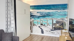 Ein Zimmer mit großem Wandbild mit Meerblick, einem grauen Sofa, einem Holztisch und einem weißen Kleiderschrank.