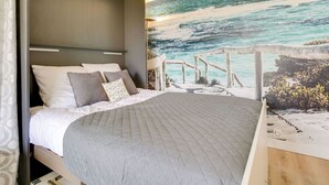 Ein Schlafzimmer mit Bett, Kopfteil, Wandbild mit Strandmotiv und einem Holztisch.