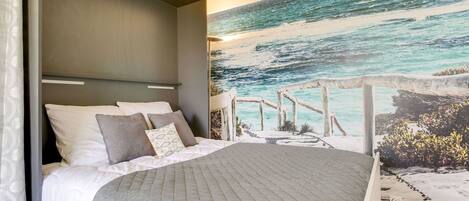 Une chambre à coucher avec un lit, une tête de lit, une fresque murale représentant une plage et un bureau en bois.