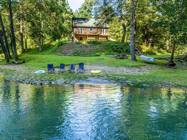 A lakeside cabin with a deck, surrounded by trees, and chairs by the water. - Secluded Cabin on a mountain stream on 7.5 acres (Lenoir)