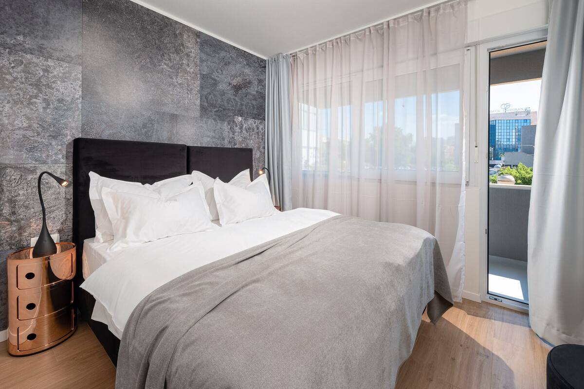 Quarto moderno com uma cama grande, mesa de cabeceira com abajur e vista da cidade através de uma grande janela.