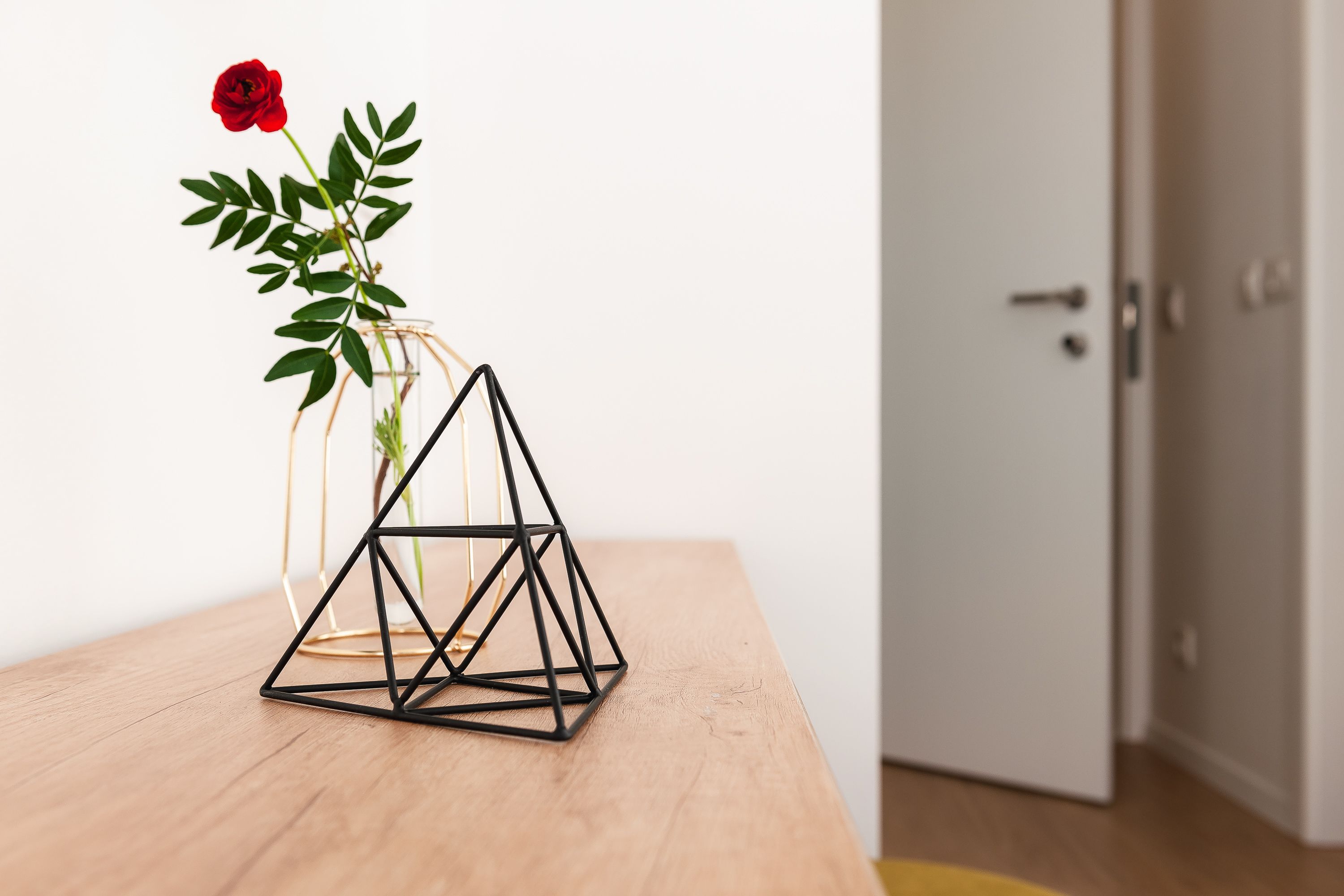 A geometric black metal sculpture on a wooden table, a vase with a red rose and green leaves, and a white door in the background.
