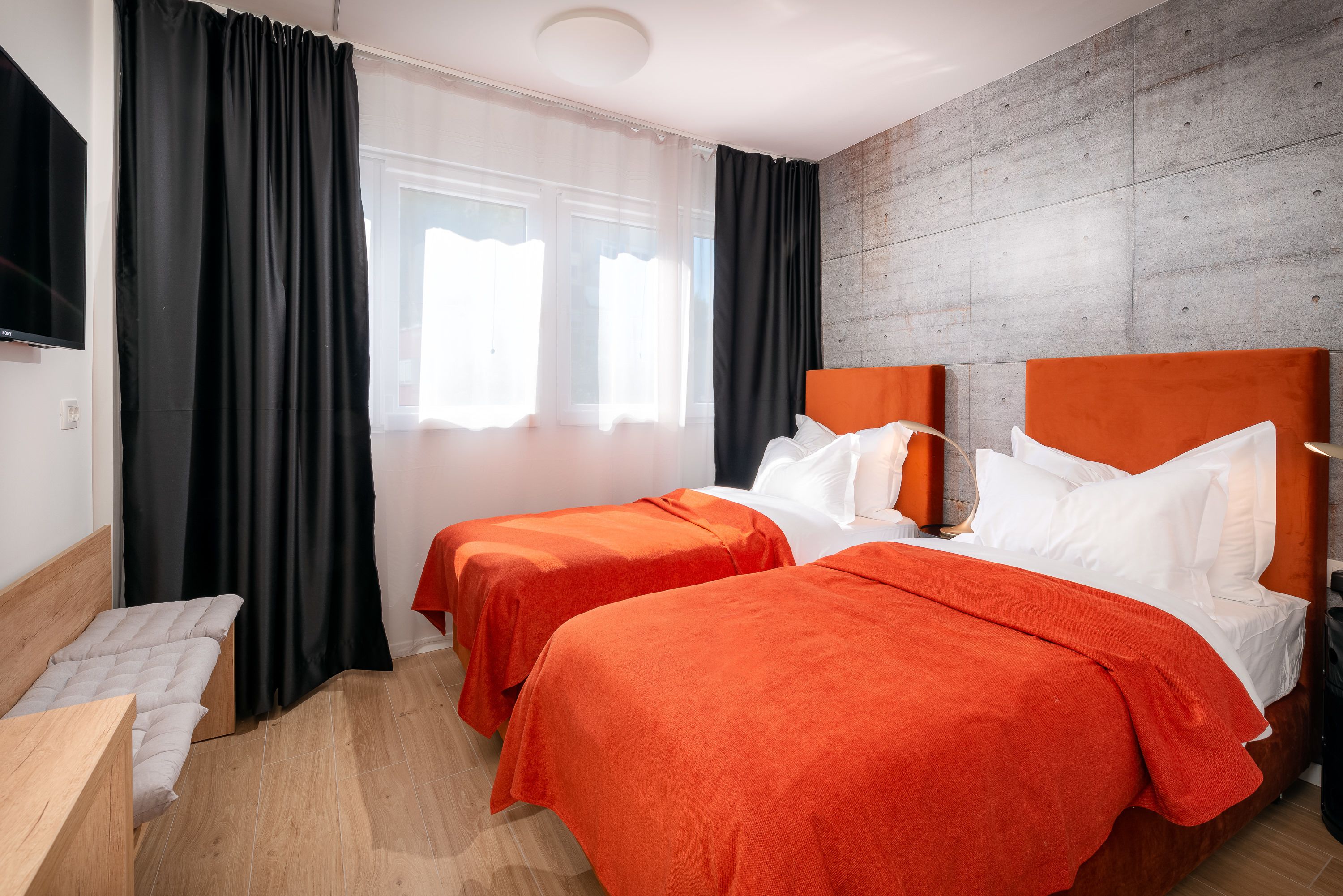 A hotel room with two beds, a bench, a flat-screen TV, and a large window with curtains.